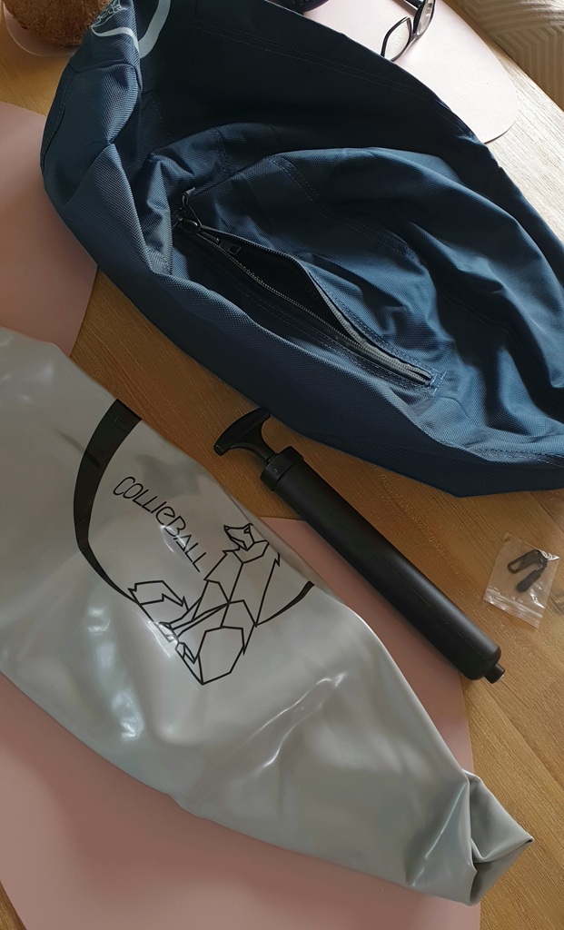 A top-down shot of the CollieBall components laid out on a timber table. It features a navy blue fabric outer shell with a zip, a deflated grey inner ball with the geometric CollieBall logo, and a black hand pump. A small plastic packet containing two black clips or pullers is also visible.