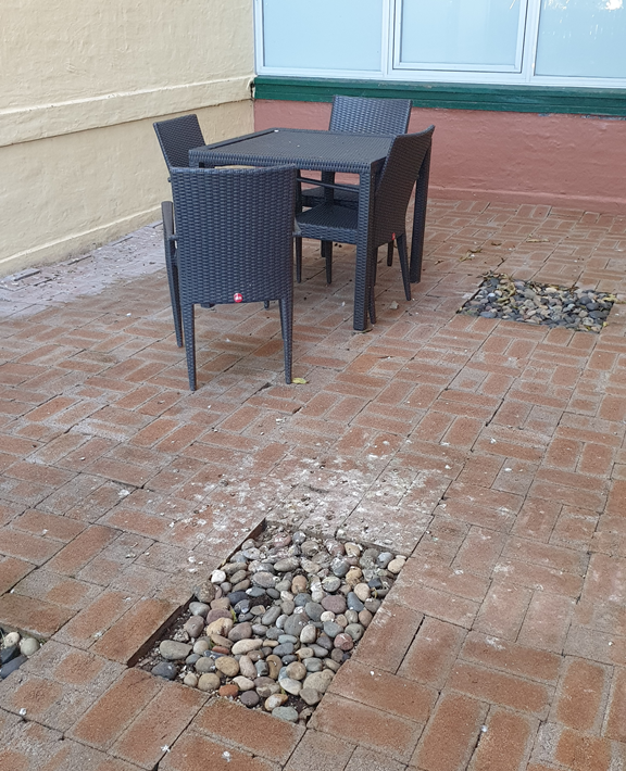 A vertical high-angle shot of a hotel courtyard paved with weathered red-brown bricks. A dark grey wicker dining set, including a square table and four chairs, sits against a cream-coloured exterior wall with large glass windows. Two rectangular sections of the paving are missing, filled instead with grey river stones and pebbles. The brickwork shows signs of white staining and scattered debris.