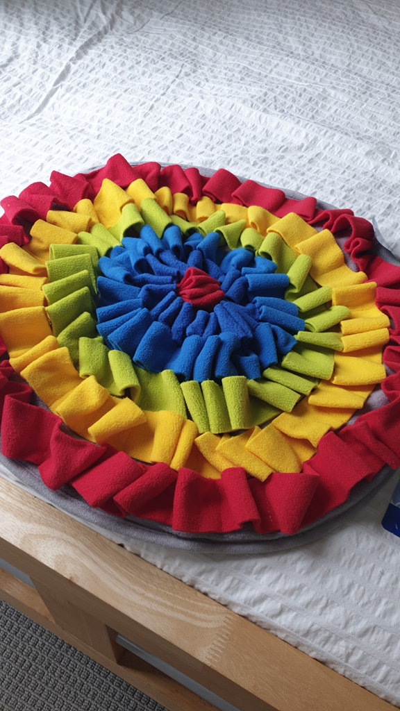 A top-down shot of a circular, multi-coloured fabric snuffle mat resting on a white textured bedsheet. The mat features concentric rings of ruffled fleece in red, yellow, green, and blue, spiralling toward a red centre. The mat is positioned on the edge of a light-coloured wooden bed frame, with a neutral grey carpet visible on the floor below.