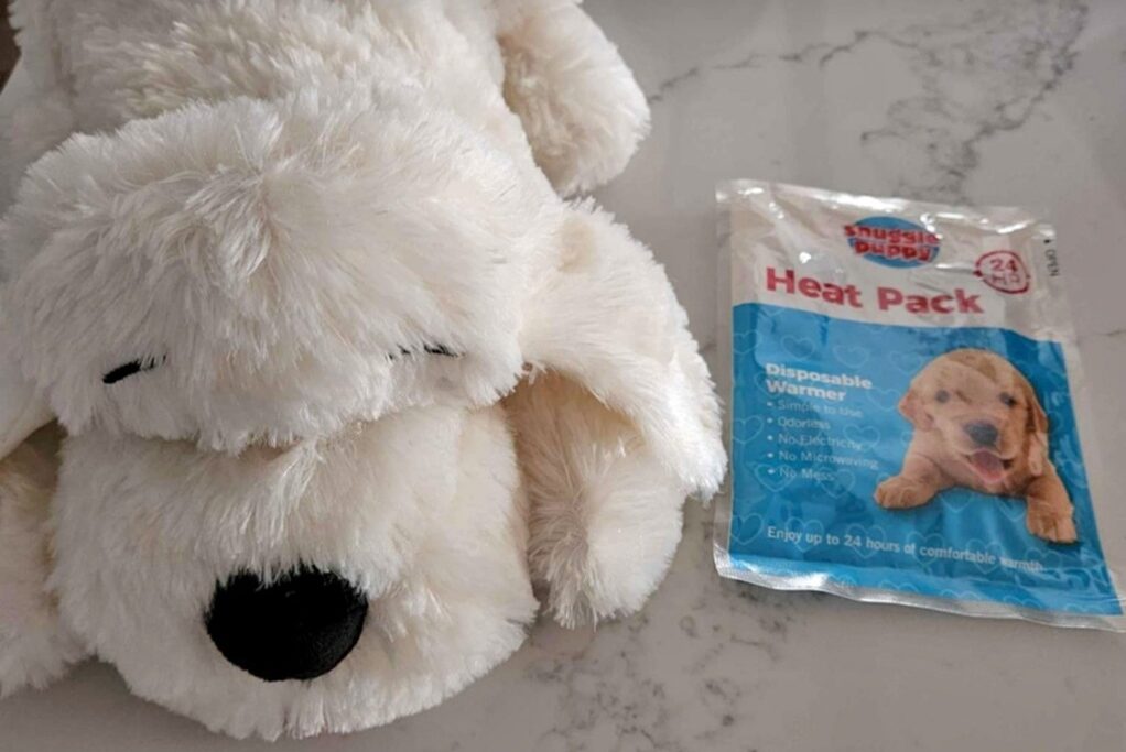 A close-up, high-angle shot of a white plush 'Snuggle Puppy' toy resting on a light-coloured marble-patterned countertop. The toy has its eyes closed and a black nose. Next to the plush toy is its original blue and white 'Heat Pack' packaging, which features a photo of a golden retriever puppy and describes it as a disposable warmer providing up to 24 hours of comfort.