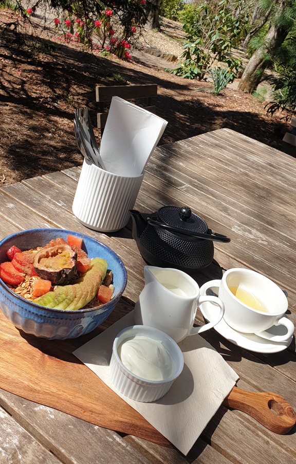 A vertical high-angle shot of a breakfast set-up on a weathered timber outdoor table. In the foreground, a blue ceramic bowl is filled with muesli topped with fresh strawberries, passionfruit, and sliced green apple. Beside it sits a small white ramekin of yoghurt on a wooden serving board, a black cast-iron teapot, a small white milk jug, and a white teacup filled with light-coloured tea. The background shows a sun-drenched garden with green trees and vibrant pink azaleas.