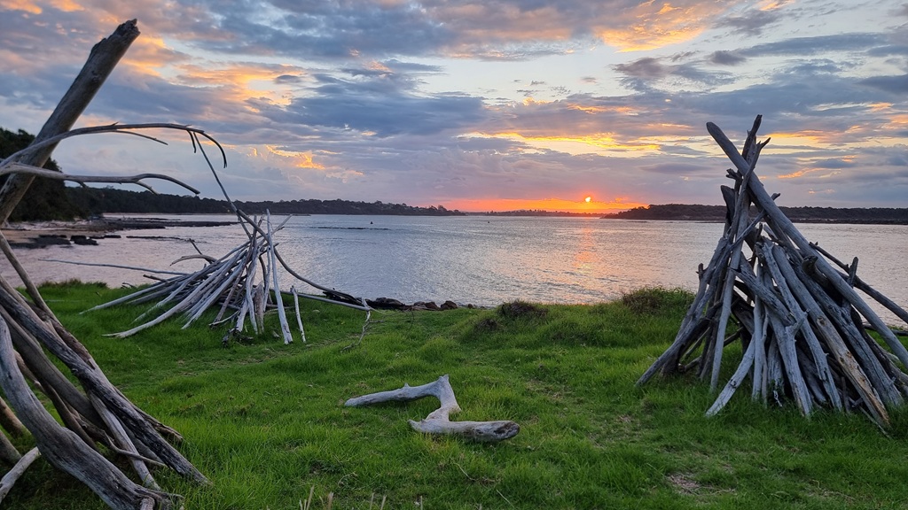 A vibrant sunset over a calm river or bay, viewed from a grassy bank. Three large teepee-style structures made of bleached driftwood branches stand on the green grass. The sun is setting on the horizon, casting orange and yellow light through a cloudy sky across the water.