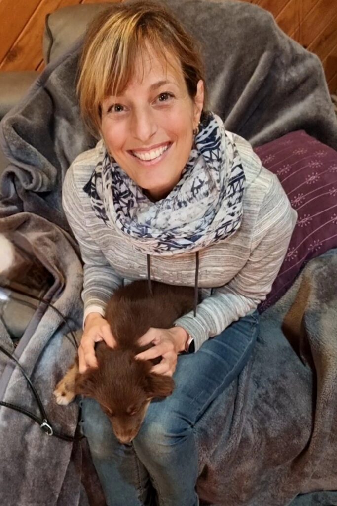 A woman with a bright smile sits in a grey plush armchair, wearing a grey striped hoodie and a blue patterned infinity scarf. She is looking directly at the camera while gently cradling a chocolate-brown Miniature American Shepherd puppy, Monte, on her lap. The puppy is resting calmly, facing away from the camera. A purple patterned pillow and a glimpse of timber wall panelling are visible in the background.