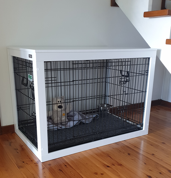 A vertical eye-level shot of a white VeboPet wooden dog crate furniture piece. The crate is positioned on polished timber floorboards under a flight of stairs. It features a white wooden frame and top with black metal wire panels. Inside the crate, a grey KONG teddy bear rests on a grey mat next to a silver metal water bowl.