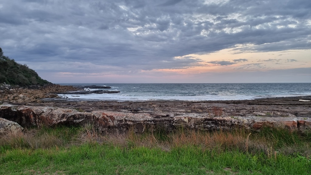 A wide landscape photograph of a rocky coastline near Crookhaven Lighthouse at dusk. In the foreground, green grass and wild shrubs sit before a low rock wall. Beyond, a dark, flat rock platform leads to the ocean under a dramatic sky filled with textured grey clouds and a soft orange sunset glow on the horizon