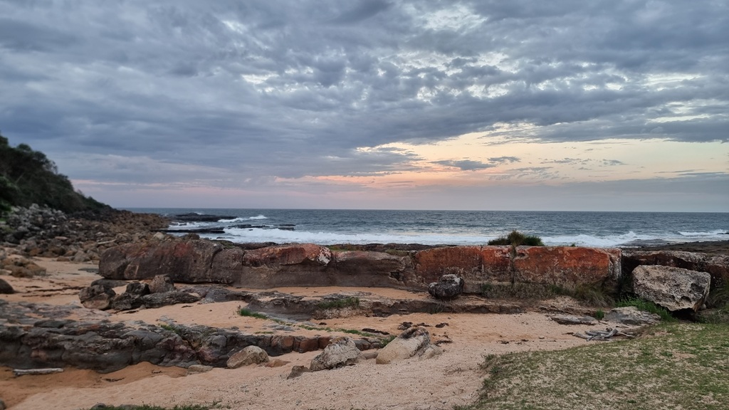 A wide landscape shot of a rocky shoreline near Crookhaven Lighthouse at dusk. Large, rust-colored boulders and flat rock platforms line the coast, meeting a churning ocean under a heavy, dramatic sky with a soft orange and pink sunset glow on the horizon.