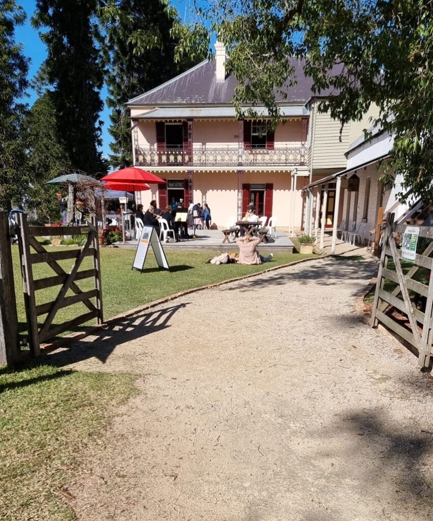 A sunny afternoon at the historic Bella Vista Farm. A gravel pathway leads through open timber gates toward the two-story pink heritage homestead where visitors relax on the lawn.