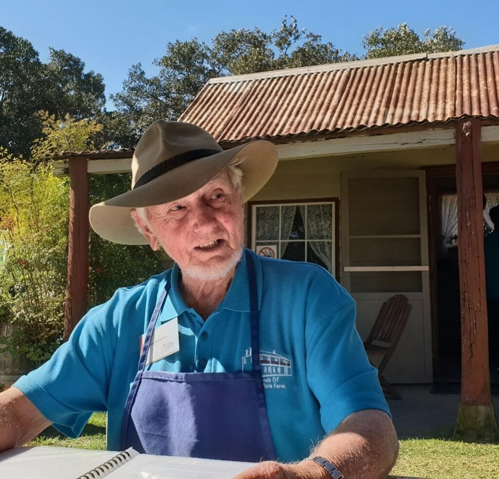 A close-up portrait of an elderly gentleman wearing a classic wide-brimmed khaki Akubra-style bush hat, a blue polo shirt featuring the "Friends of Bella Vista Farm" logo, and a dark blue apron. He is sitting outdoors on a sunny day at a table with an open binder, looking off-camera with an engaging expression. In the background is a rustic, weathered cottage with a corrugated iron roof and timber pillars.