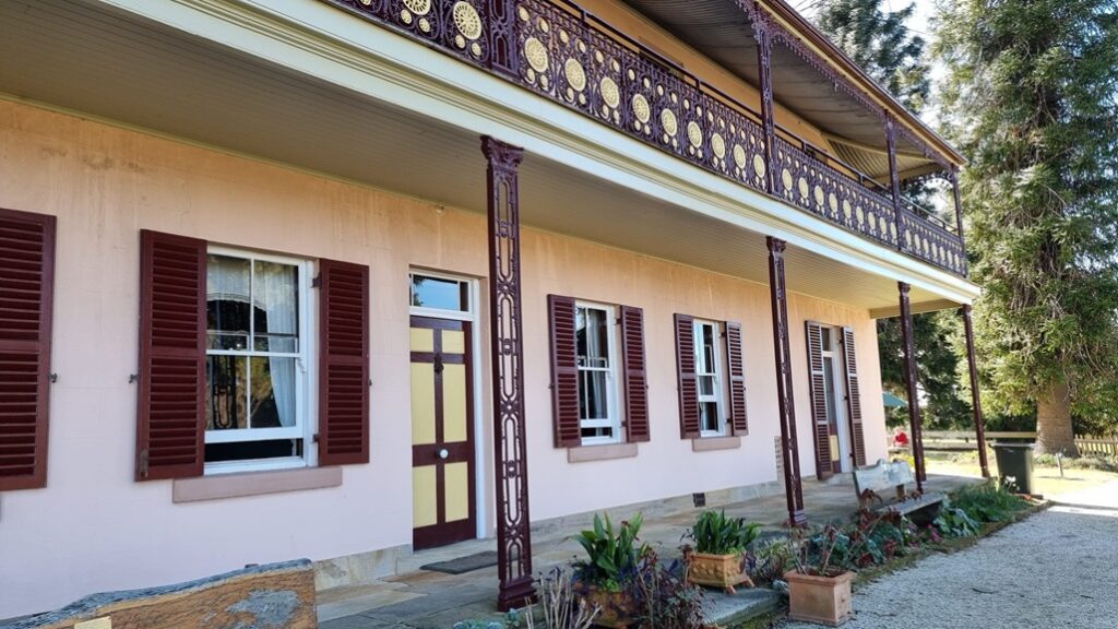 A close-up view of the historic Bella Vista Farm homestead. The pink sandstone facade is lined with multiple dark red shuttered windows and an elegant cream and maroon front door. Ornate cast-iron lace columns support the upper-story balcony, which features detailed maroon and gold iron railings. The sandstone veranda is edged with potted plants and wooden bench seating, capturing the textures of the early colonial architecture.