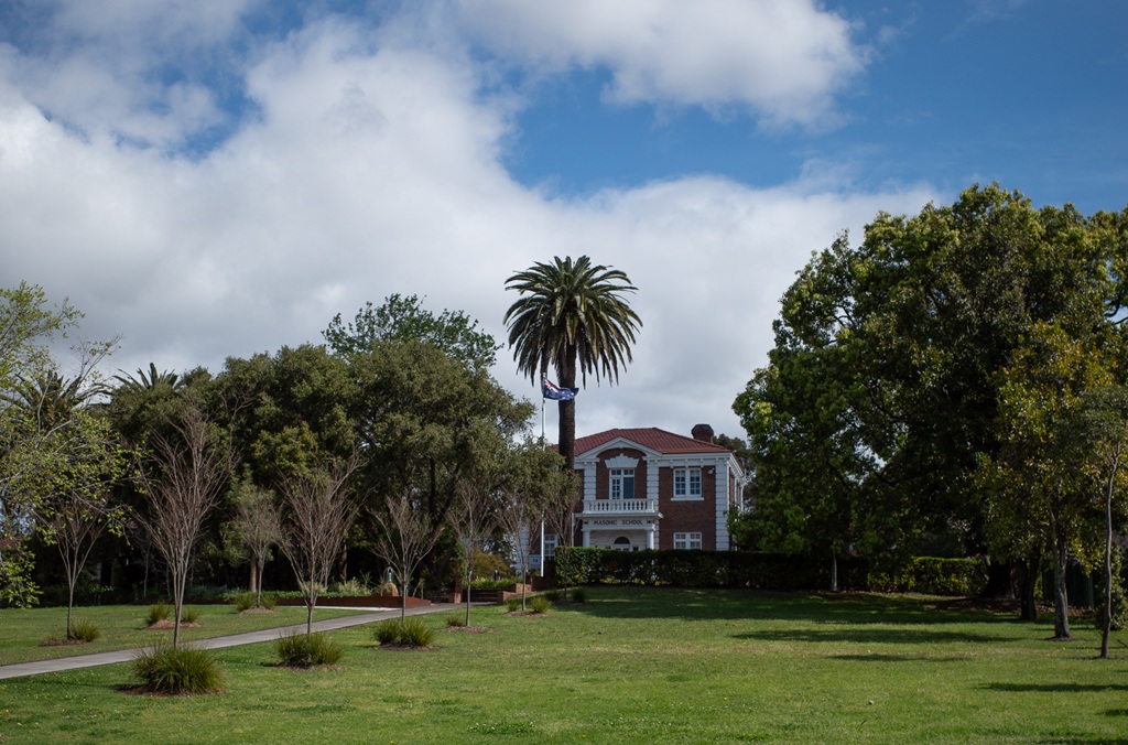 A historic two-story brick heritage building with white balconies stands behind a green lawn and mature trees under a blue sky with white clouds.