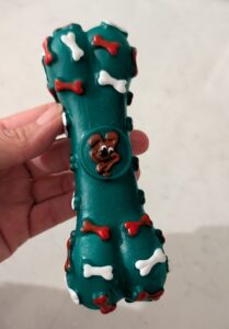 A hand holding up a dark green rubber bone-shaped dog toy featuring a cartoon dog face in the centre and small raised red and white bone shapes.