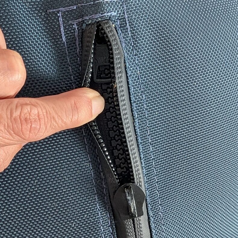 A finger peeling back the protective fabric flap of a dog herding ball to reveal the heavy-duty black plastic zipper tracks and the inner zipper pull slider beneath.