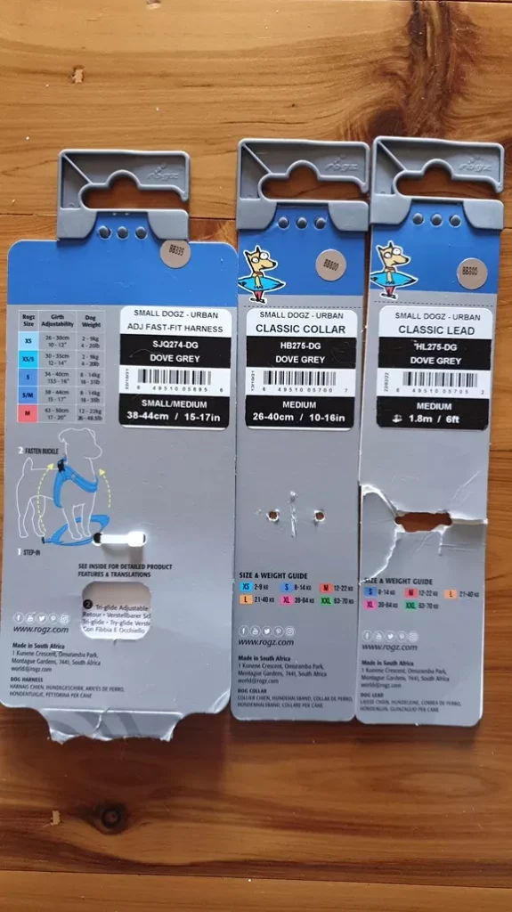 Three Rogz Urban product cardboard packaging tags displayed on a wooden table, showing details for the Fast-Fit Harness, Classic Collar, and Classic Lead in Dove Grey.