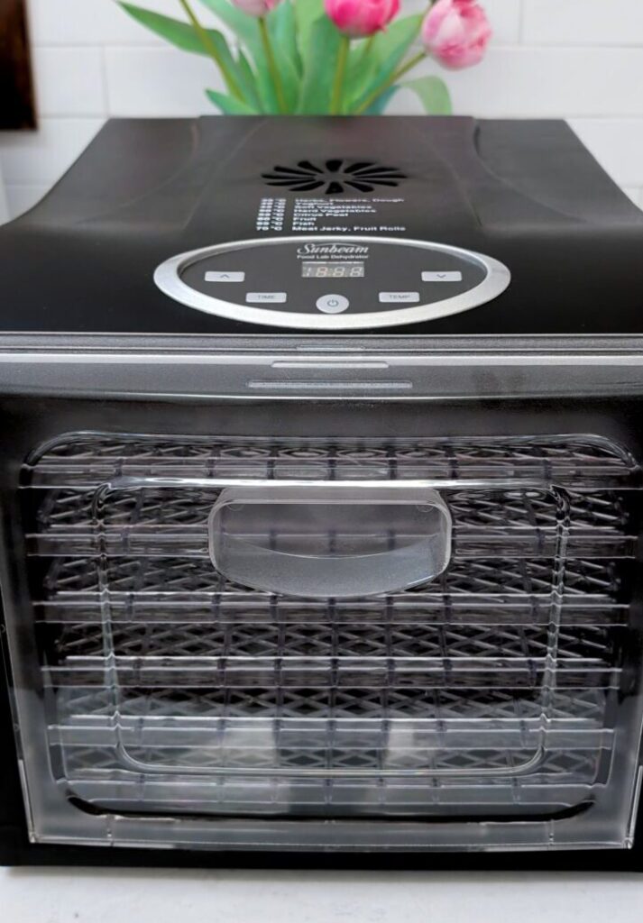 A black Sunbeam Food Lab multi-tray food dehydrator with a digital display, sitting on a white kitchen benchtop with a vase of pink tulips in the background.