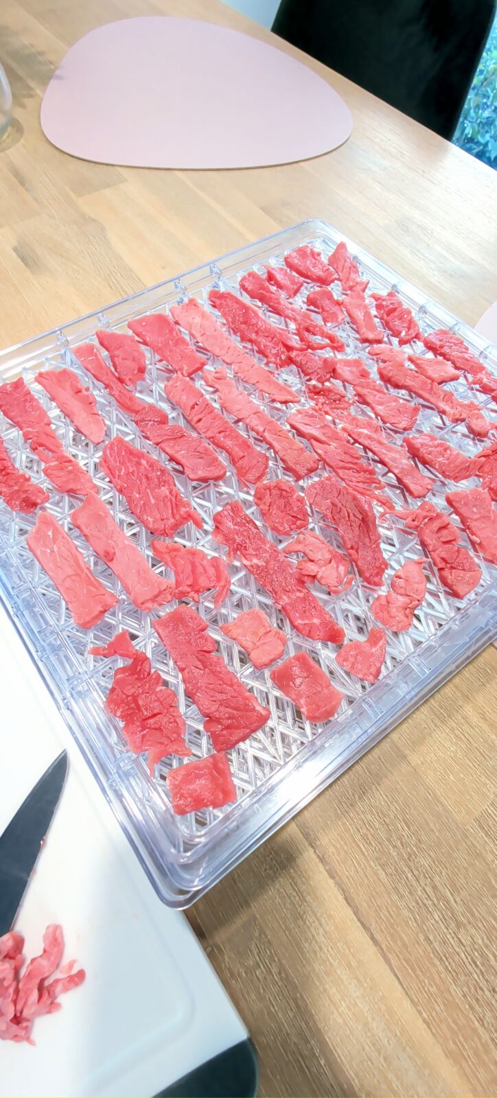 Thinly sliced raw beef strips arranged evenly on clear dehydrator trays resting on a light wooden table.