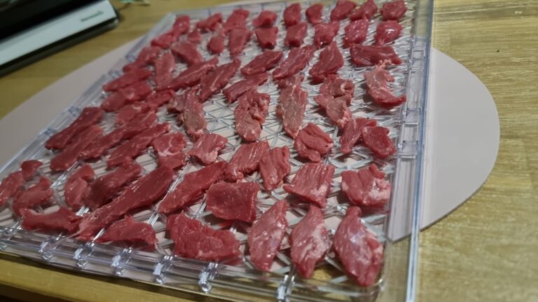 Thinly sliced raw red meat arranged evenly on clear dehydrator trays resting on a light wooden table, with a knife and cutting board visible in the corner.