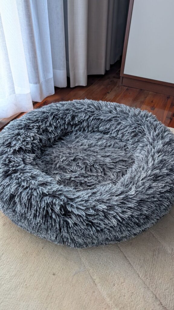 A side-angle view of a circular, dark grey shaggy faux-fur Pawz calming donut dog bed sitting on a light-coloured rug next to a polished timber floor and light curtains, showcasing its plush, fluffy texture.