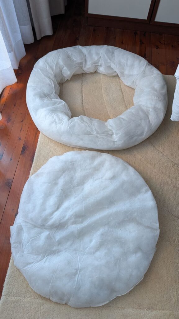 A top-down view of the two white fabric internal cushion inserts removed from a round Pawz calming donut dog bed, showing the large ring bolster piece and the flat circular centre cushion laying separate on a light rug over timber floors.