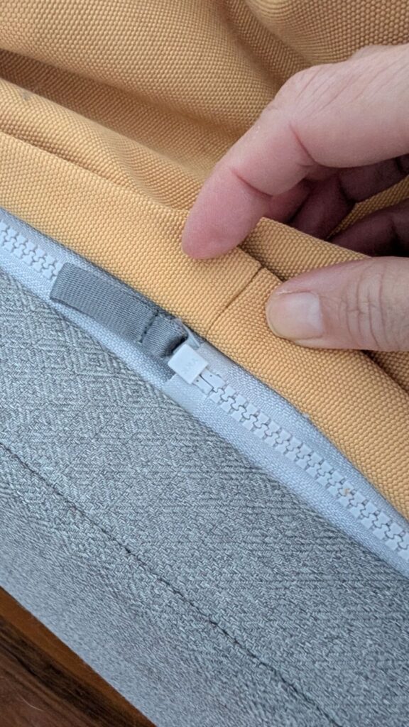 A close-up view of fingers pressing down a thick yellow canvas fabric flap on an Omlet Topology dog bed to demonstrate how it completely covers and conceals the grey zipper track system connecting the topper to the base.