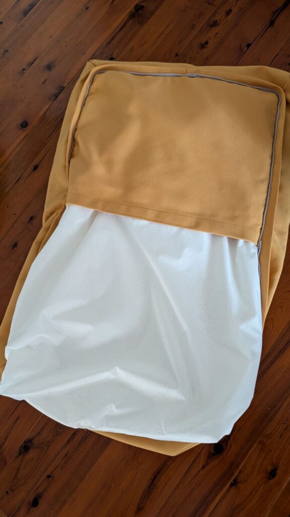 A close-up view of the underside of a yellow Omlet Topology dog bed beanbag topper cushion with the zipper undone, revealing a white inner lining fabric bag holding the beans being partially pulled out on a timber floor.