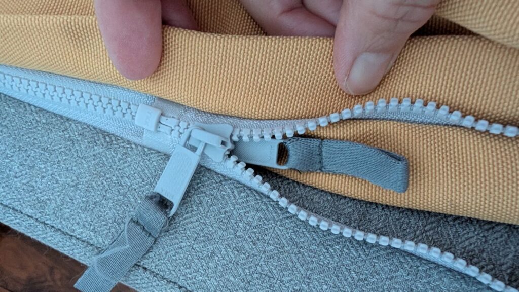 A close-up view of a hand lifting a thick fabric flap on a yellow Omlet Topology dog bed topper to reveal a heavy-duty light grey zipper tracking system attaching it to the grey base, showing how the zipper pull tabs are tucked out of sight.