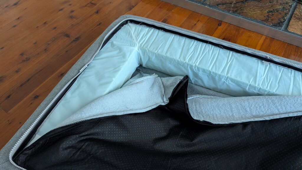 A close-up view of the underside of a grey Omlet Topology bolster dog bed topper unzipped, revealing the internal L-shaped white foam bolster cushion insert being accessed for removal on a timber floor.