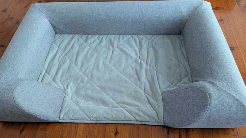 A top-down view of a grey fabric Omlet Topology bolster dog bed topper resting directly on polished timber flooring, featuring raised padded bolster sides and a quilted pale green central lining base.