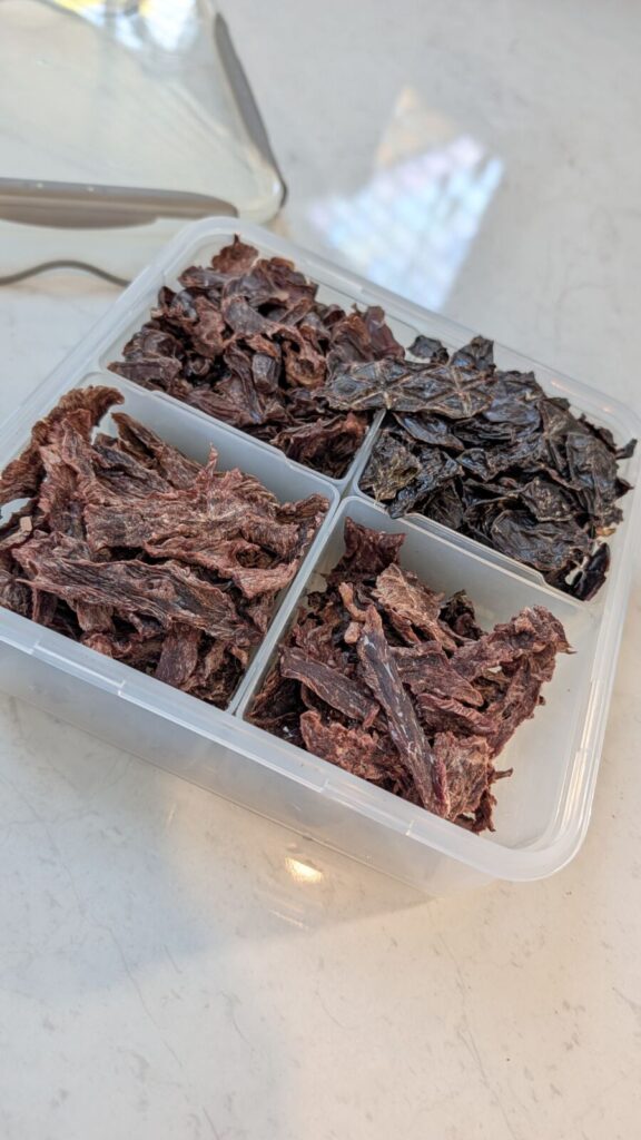 A clear plastic storage container divided into four sections, each filled with a different type of dark, homemade dehydrated meat treat, sitting on a white kitchen bench.