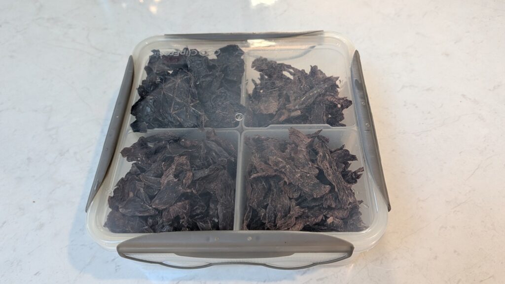 A square, clear plastic storage container with its lid securely clipped on, divided into four sections filled with dark, homemade dehydrated meat treats, sitting on a white kitchen bench.