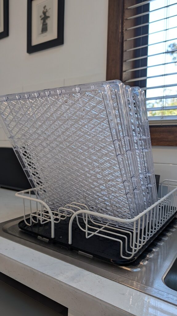 Clear plastic dehydrator trays stacked vertically to air dry in a white wire dish rack next to a kitchen sink.