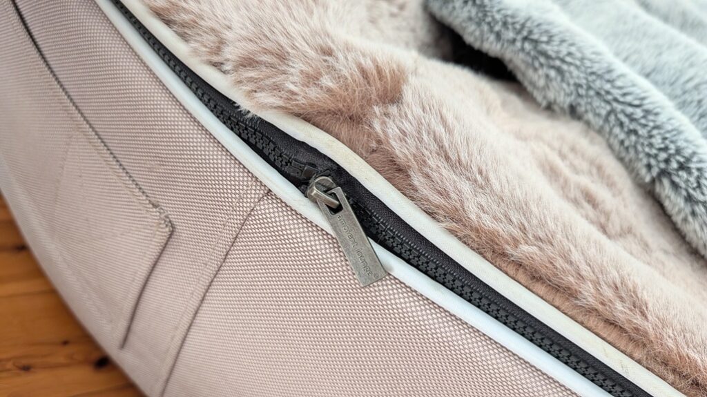 A close-up, side-angle shot of the metal zipper pull on an Ambient Lounge dog bed, showing the rectangular puller lying flat against the beige woven fabric base right next to the plush faux-fur cappuccino topper.