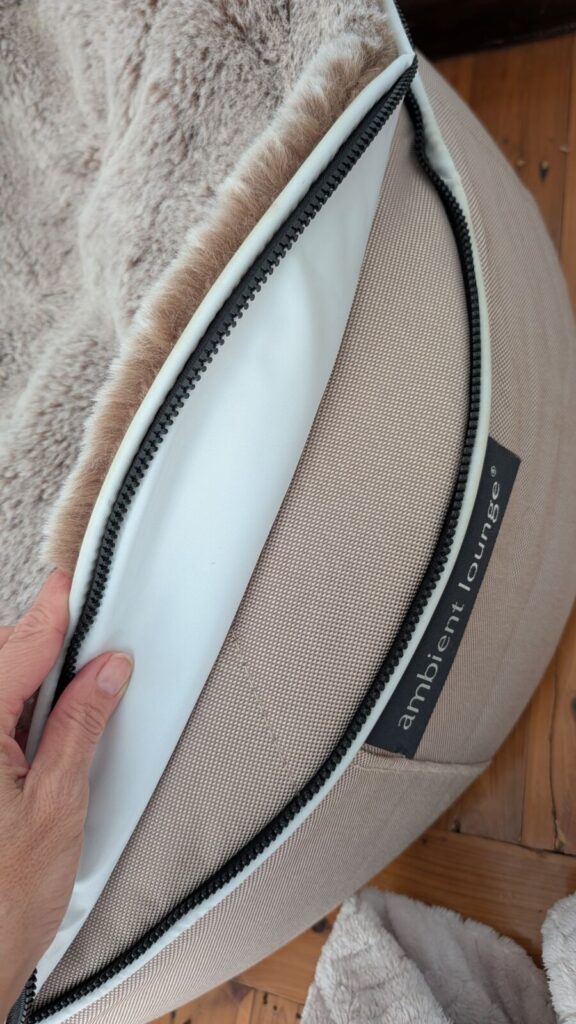 A close-up view of the black zipper track on a beige Ambient Lounge dog bed, showing a hand lifting the plush faux-fur cappuccino topper to reveal the robust zipper attachment along the heavy-duty base fabric.