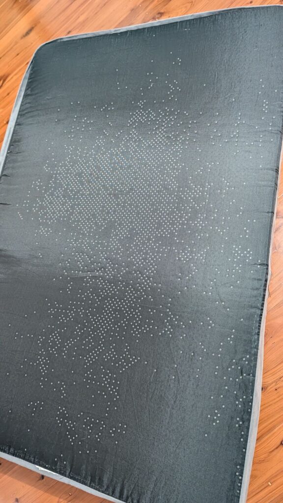 The black non-slip base of an orthopaedic dog bed mattress layout on a timber floor, showing hundreds of tiny rubber grip dots peeling and washing off after being laundered.