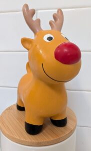 A close-up shot of a yellow latex reindeer dog toy with large antlers, a big red nose, and a painted smile, standing on a round bamboo lid against a white tiled wall.