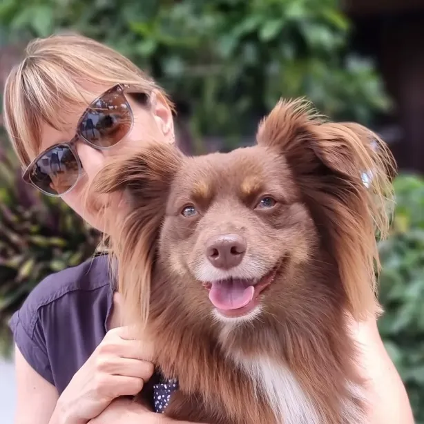 A close-up outdoor portrait of Caroline smiling and holding her fluffy brown Mini American Shepherd, Monte. Caroline is wearing sunglasses and a dark grey top. Monte is in the foreground, looking directly at the camera with a happy, open-mouthed expression, showcasing his distinctive golden-brown fur and white chest markings against a softly blurred green foliage background.