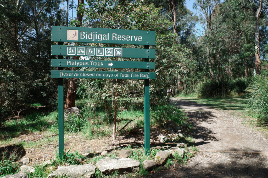 A green timber park entrance sign for Bidjigal Reserve and the Platypus Track pointing towards a dirt bush path.