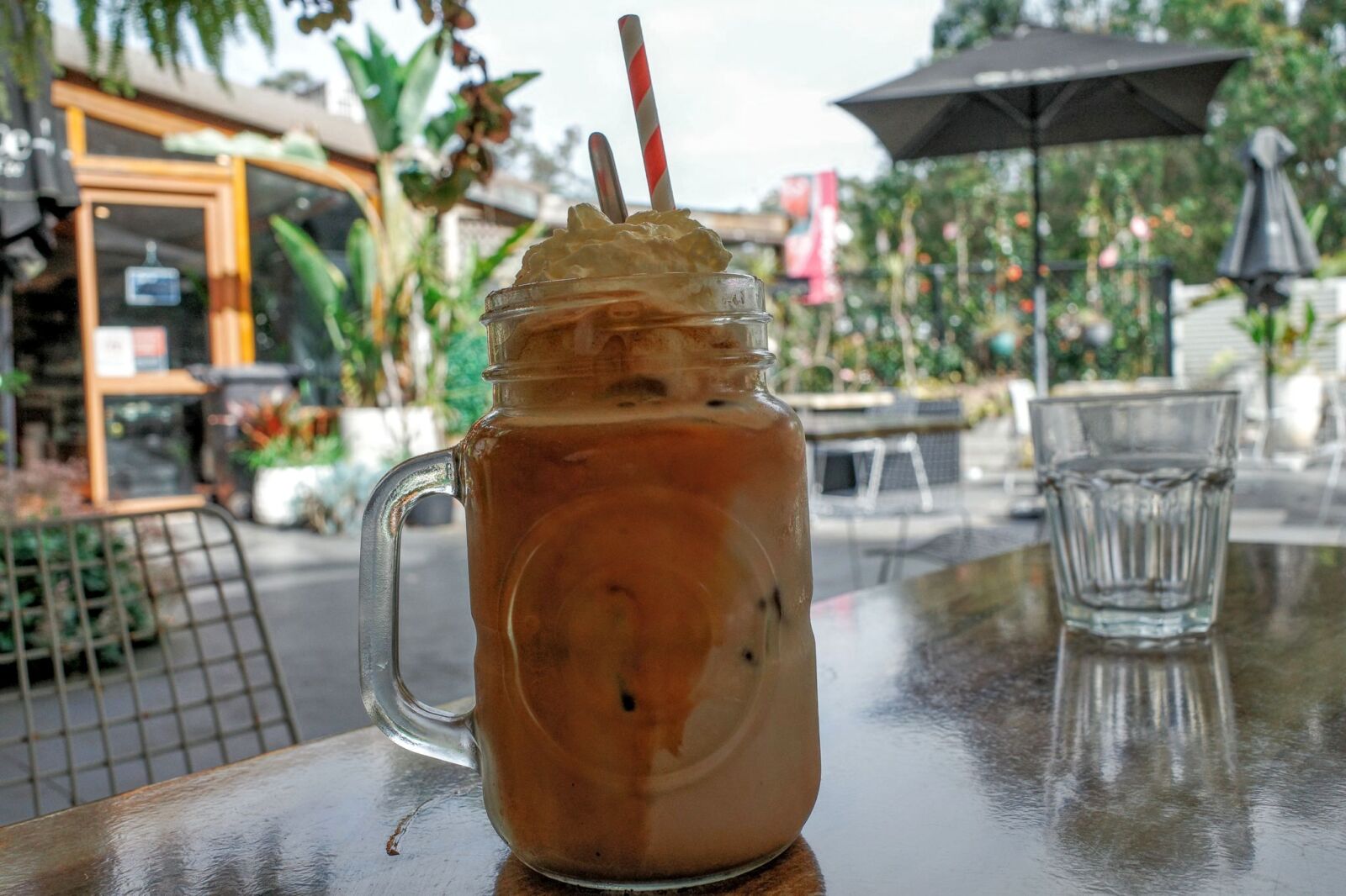 An iced coffee served in a glass mason jar mug topped with whipped cream and two striped paper straws sits on an outdoor wooden table at Café Saligna, with the café building and empty outdoor terrace visible in the blurred background.