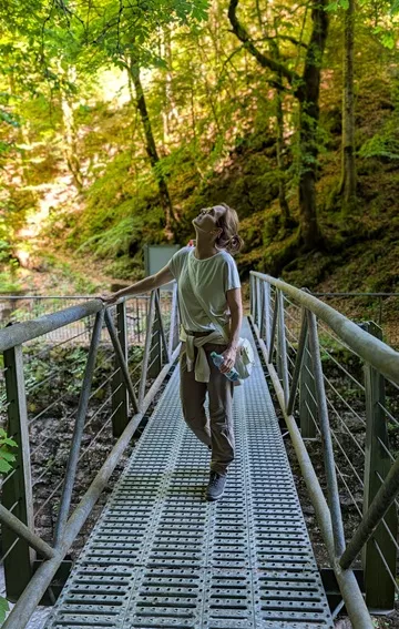 Woman wearing an ivory Wag Society Memory Walk Pouch and hiking pants, holding a water bottle, and looking up while crossing a metal grate suspension bridge over an Alpine gorge.