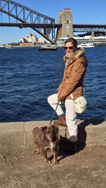 Woman wearing a brown puffer jacket, sunglasses, and the ivory Wag Society Memory Walk Pouch crossbody while standing with her Miniature American Shepherd by Sydney Harbour, with the Sydney Opera House and Harbour Bridge in the background.