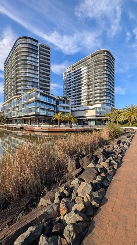 A paved brick pathway lined with dark rocks and dry reeds alongside the lake, looking toward modern high-rise buildings and palm trees under a blue sky.