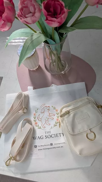 Flat lay of the ivory Wag Society Memory Walk Pouch, two interchangeable straps, and branded packaging next to a vase of pink tulips.