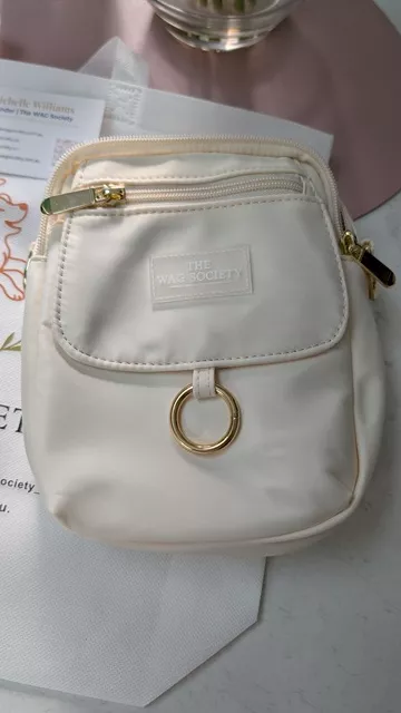 Front view of the ivory Wag Society Memory Walk Pouch, showcasing its clean nylon fabric, front zippered pocket, branded label, and gold hardware ring.