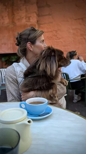 Caroline holding her brown Miniature American Shepherd Monte close while sitting at a marble cafe table with tea in the courtyard.