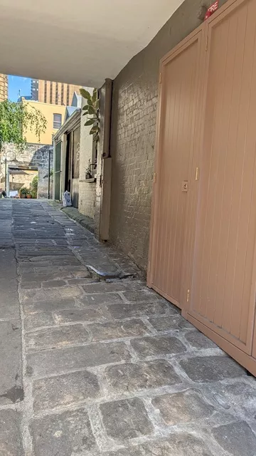 A narrow stone-paved alleyway beside a building with beige doors, leading toward an open courtyard in The Rocks.
