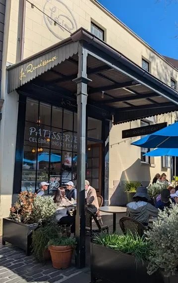 The sunny exterior storefront of La Renaissance Patisserie in The Rocks, featuring outdoor seating, blue umbrellas, and potted plants.
