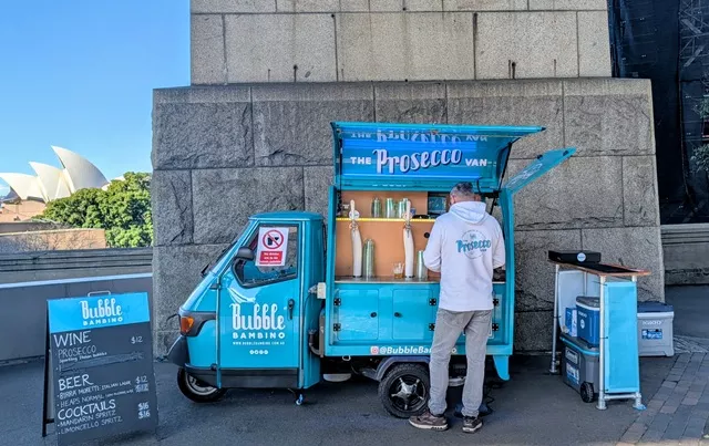 A bright turquoise mobile Prosecco van named Bubble Bambino parked outdoors near Sydney Harbour, with a menu board displaying wine, beer, and cocktails.