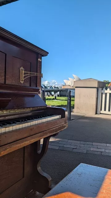 An upright wooden piano placed outdoors with the Sydney Opera House visible in the background under a clear blue sky.