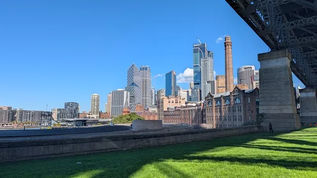A wide view of the Sydney city skyline and historic brick buildings captured from grassy parkland near the Sydney Harbour Bridge.