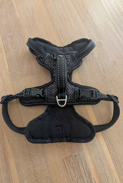 Top-down flat lay view of the black Rogz Utility Explore dog harness on a wooden floor, highlighting the top control handle, side-release buckles, and sturdy D-ring.