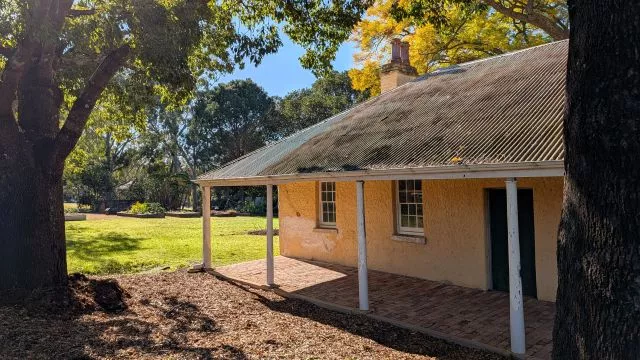 A historic colonial-era stone cottage with a corrugated iron roof and veranda, nestled among trees and lawns at Parramatta Park.